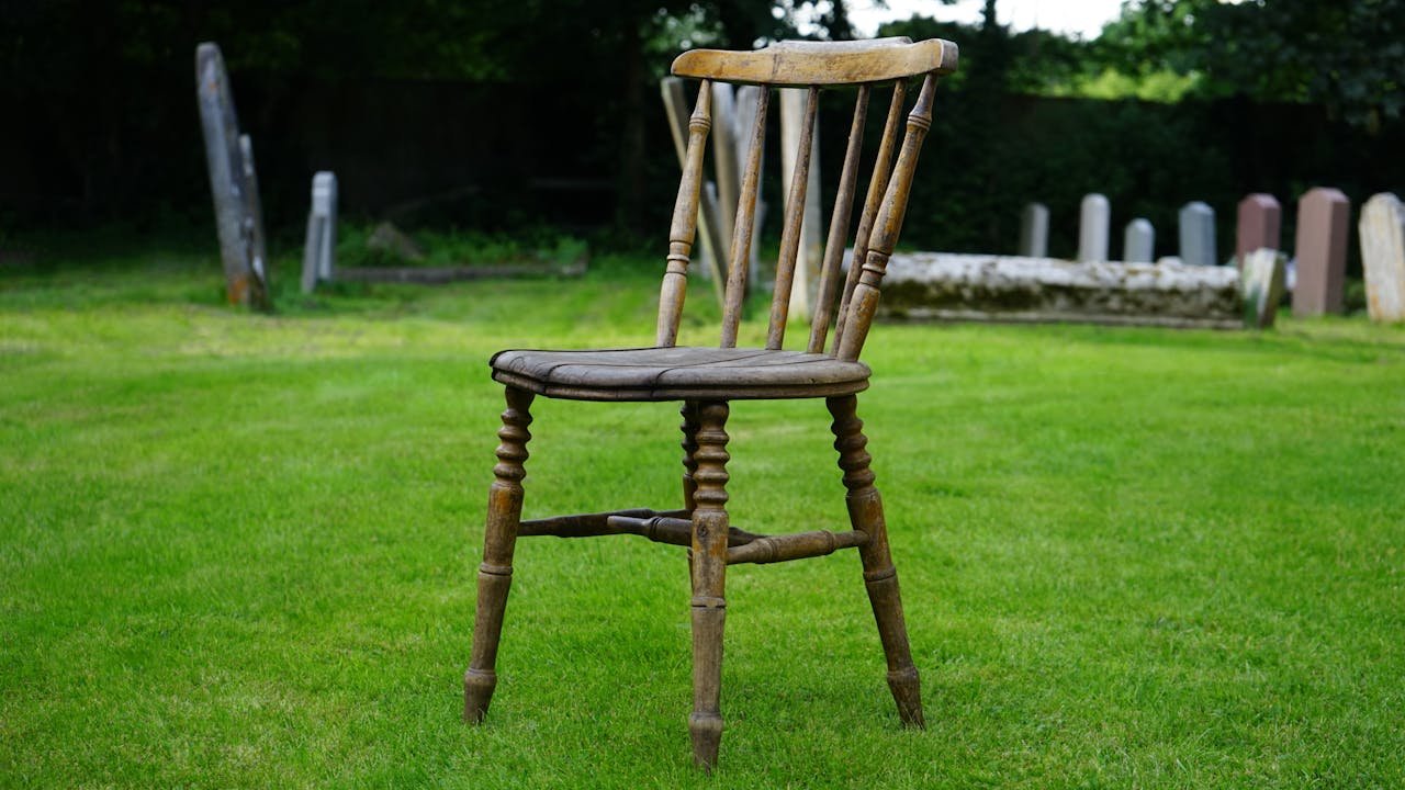 A vintage wooden chair sits alone in a serene cemetery surrounded by tombstones.